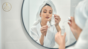 Woman with sensitive skin applying a gentle luxury skincare serum in front of a mirror, wearing a towel and robe during a calm self-care routine.